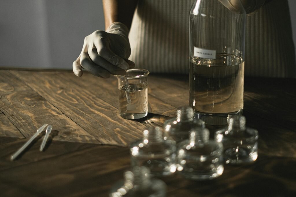 Crop anonymous female in latex gloves making fluid for perfume among glass jars on wooden table